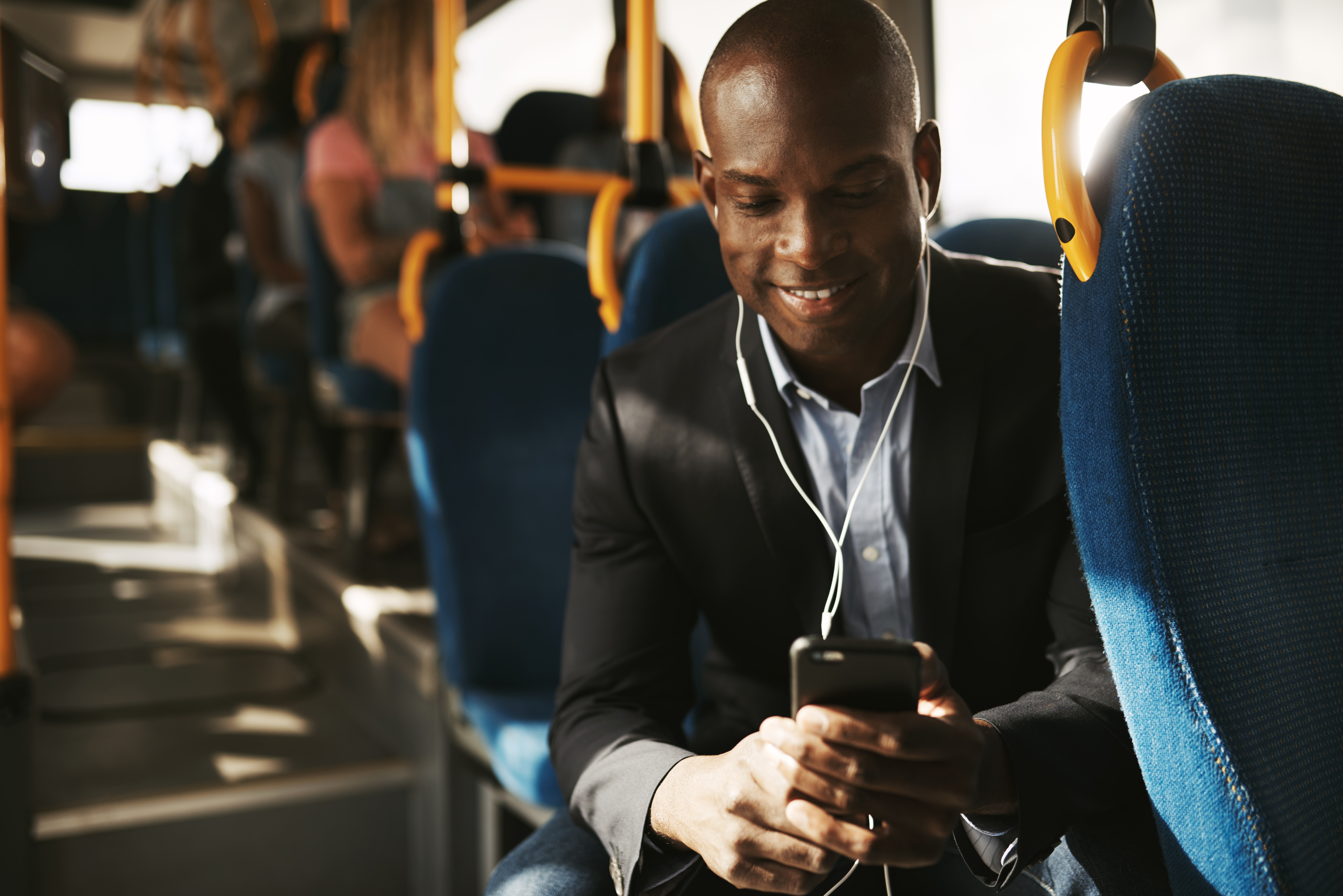 Smiling young African businessman wearing a suit sitting on a bus during his morning commute listening to music on a smartphone and earphones